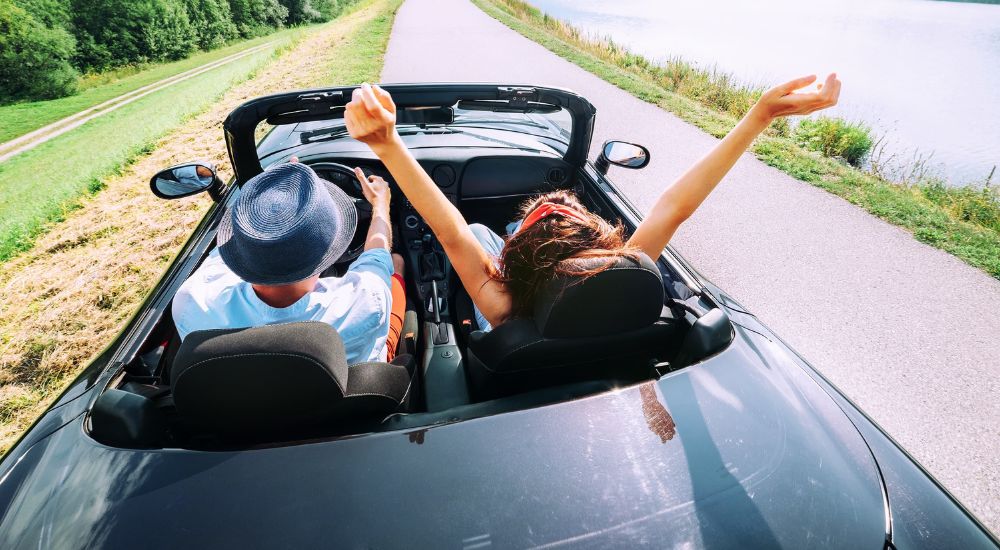 A couple are shown sitting in a convertible driving down a road by the ocean on a sunny day.