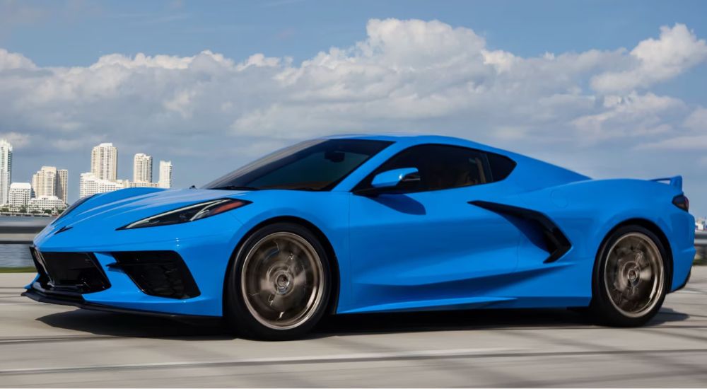 A blue 2023 Chevy Corvette is shown driving down an open highway in front of a city skyline.
