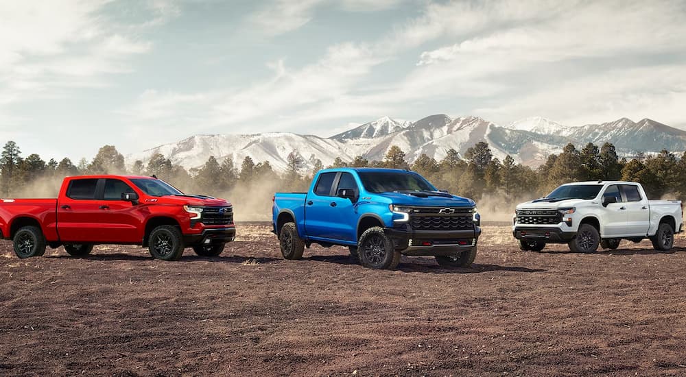 Three 2023 Chevy Silverado 1500s, available at a Chevy truck dealer, are shown parked in the dirt with mountains in the background.
