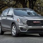 A gray 2023 GMC Terrain for sale is show parked in front of trees.