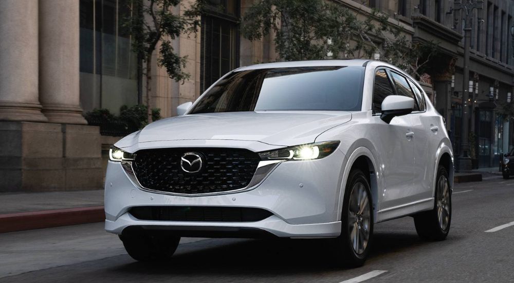 A white 2023 Mazda CX-5 is shown driving through the city.
