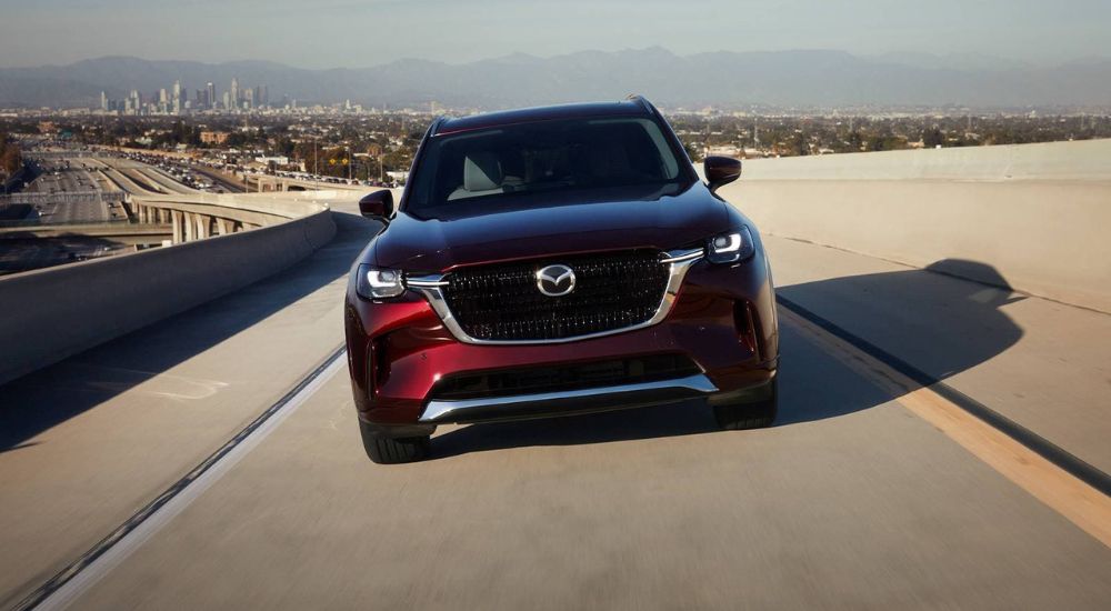 A red 2024 Mazda CX-90 is shown driving directly toward the camera on a highway.