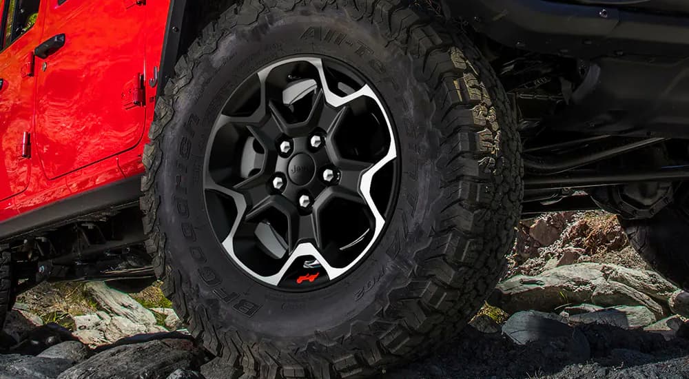 An all-terrain tire on a red 2023 Jeep Wrangler Rubicon is shown traversing rocky ground.