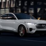 A white 2023 Ford Mustang Mach-E is shown driving near a building.