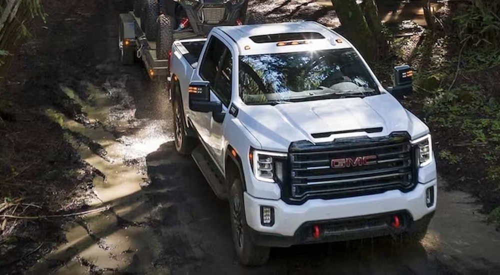 A white 2023 GMC Sierra 2500 HD AT4 is shown towing a trailer on a muddy path.