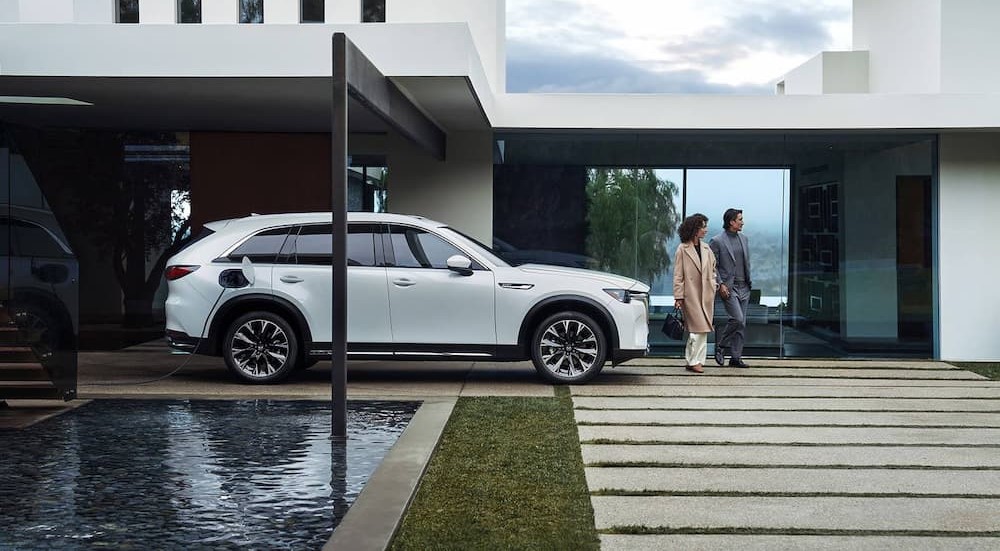 A white 2024 Mazda CX-90 PHEV is shown charging near a house.