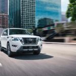 One of the most popular Nissan SUVs, a white 2023 Nissan Armada, is shown driving near city buildings.