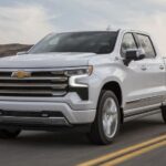 A white 2024 Chevy Silverado 1500 High Country is shown driving on a highway.