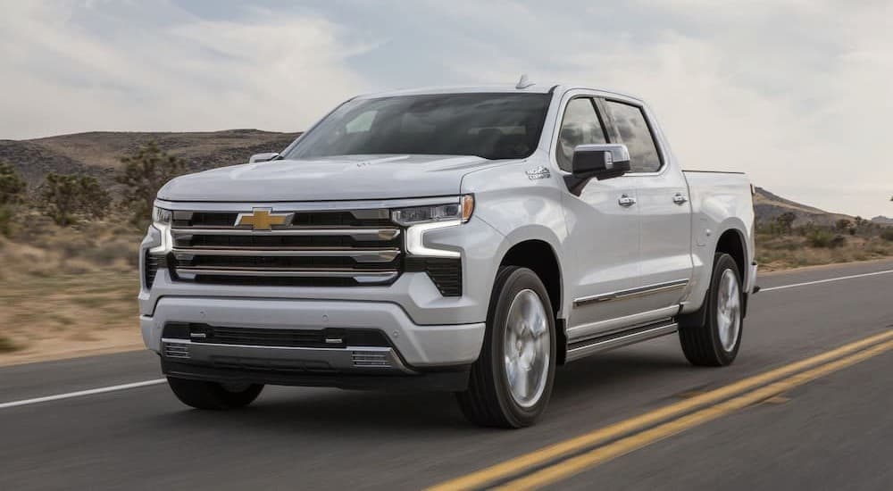 A white 2024 Chevy Silverado 1500 High Country is shown driving on a highway.