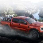 A red 2024 Ford Ranger is shown driving off-road near a river towards the debate of a 2024 Ford Ranger vs 2024 Toyota Tacoma.