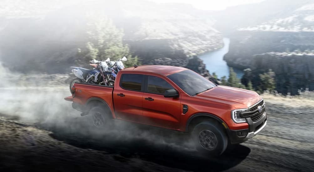 A red 2024 Ford Ranger is shown driving off-road near a river towards the debate of a 2024 Ford Ranger vs 2024 Toyota Tacoma.