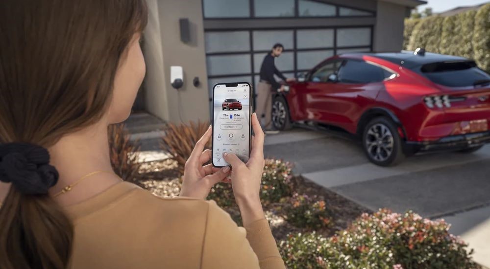 A person is checking the status of a red 2021 Ford Mustang Mach-E charging on a driveway.