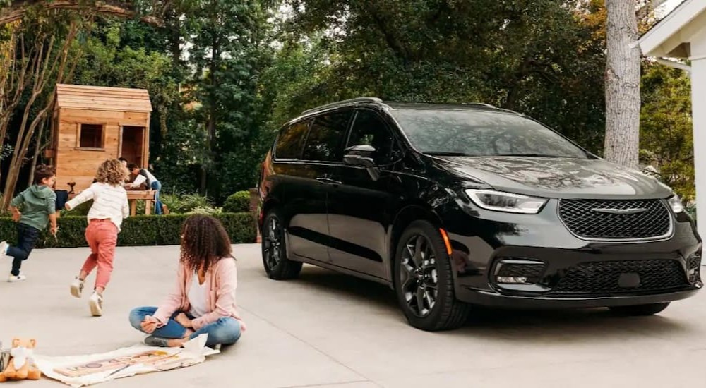 A black 2023 Chrysler Pacifica for sale is shown parked on a driveway near a family.