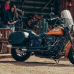 A black and orange 2023 Harley-Davidson Heritage Classic is shown parked near a wooden cabin.