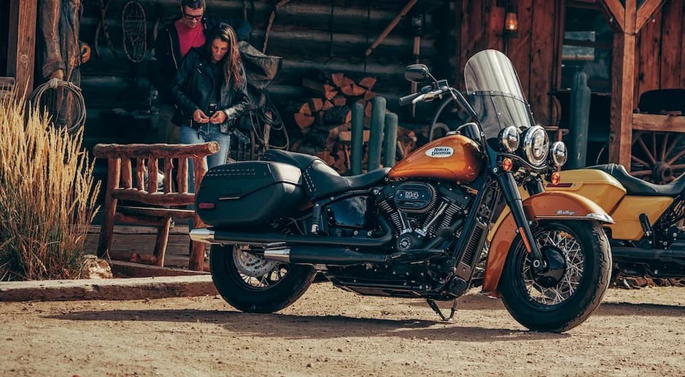 A black and orange 2023 Harley-Davidson Heritage Classic is shown parked near a wooden cabin.