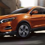 An orange 2022 Nissan Rogue Sport is shown driving on a city street after viewing a Nissan Rogue for sale.