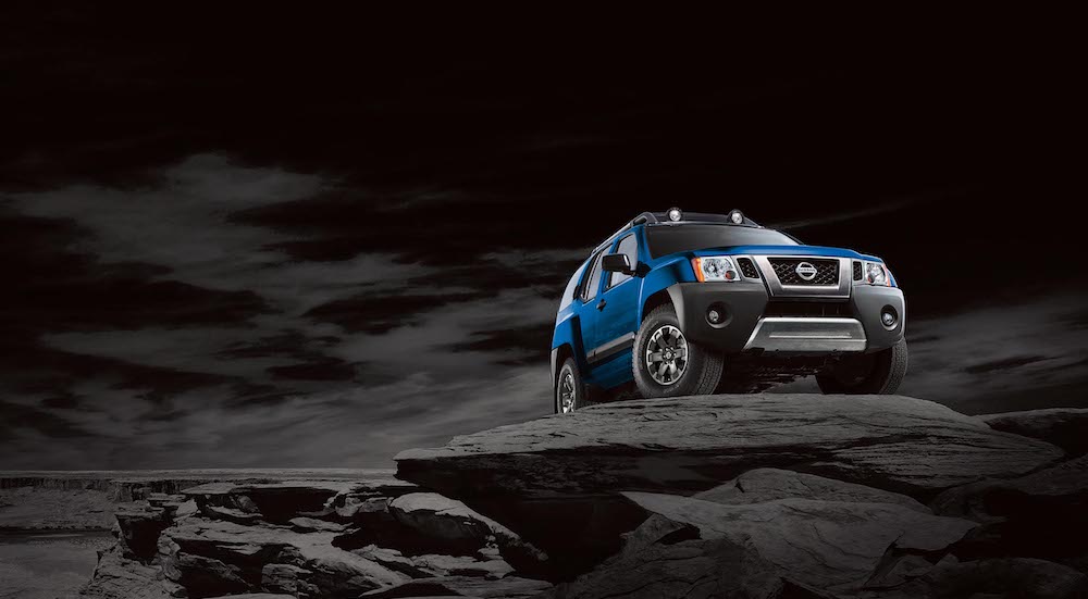 One of the older used SUVs for sale, a blue 2015 Nissan Xterra, is shown parked on a dark rocky cliff.