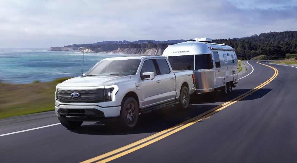 A white 2023 Ford F-150 Lightning is shown towing a trailer by the ocean.