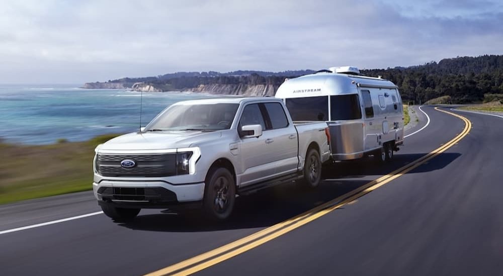 A white 2023 Ford F-150 Lightning is shown towing a trailer near an ocean.