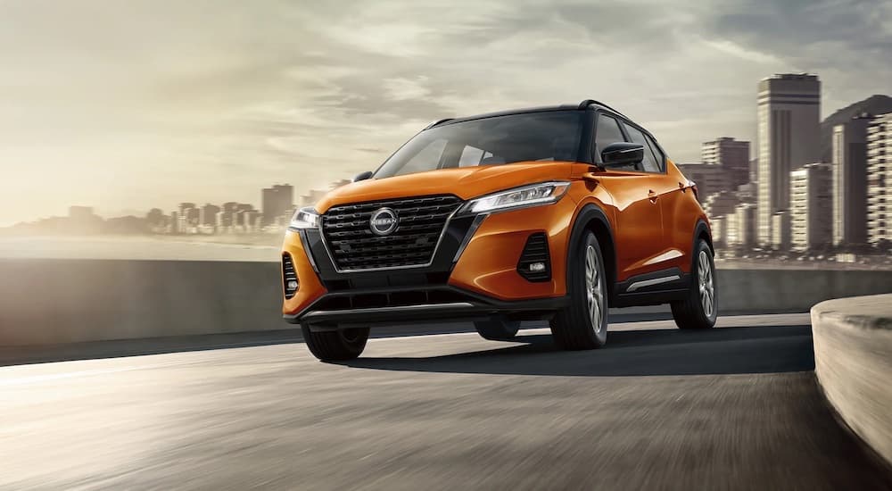 An orange 2023 Nissan Kicks S is shown driving on a bridge with a cityscape in the background.