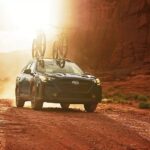 A blue 2024 Subaru Outback is shown with bikes on the roof driving on a trail.
