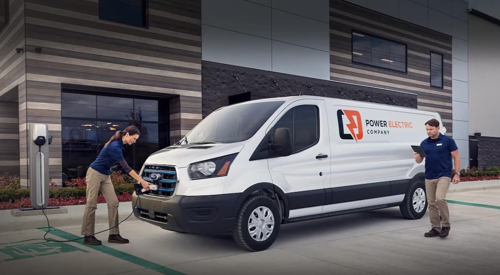 A white 2023 Ford E-Transit van is shown parked near a building and being charged.