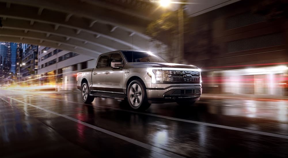 A silver 2024 Ford F-150 Lightning is shown driving on a city street near a bridge.