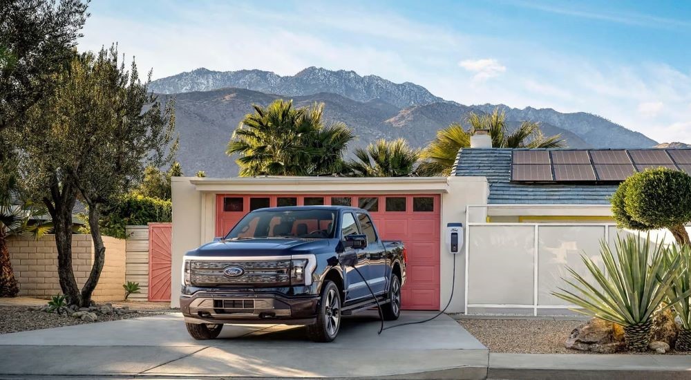 A blue 2022 Ford F-150 Lightning plugged in to charge at a modern home in the desert.