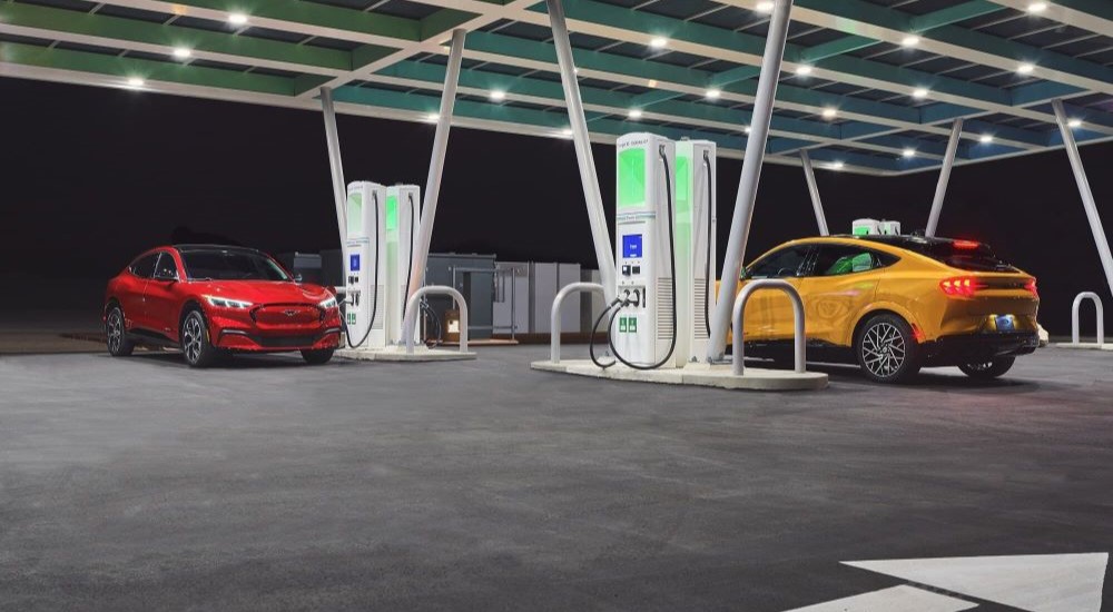 Two 2023 Ford Mustang Mach-Es, one red and one yellow, parked at a brightly lit EV charging station.