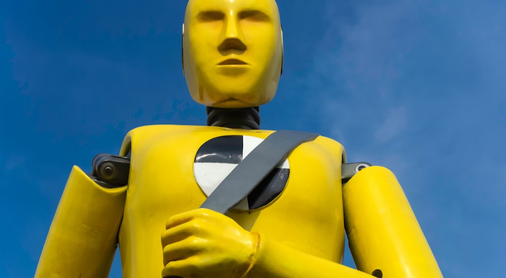 A yellow crash dummy is shown holding his seatbelt near a used car dealer.