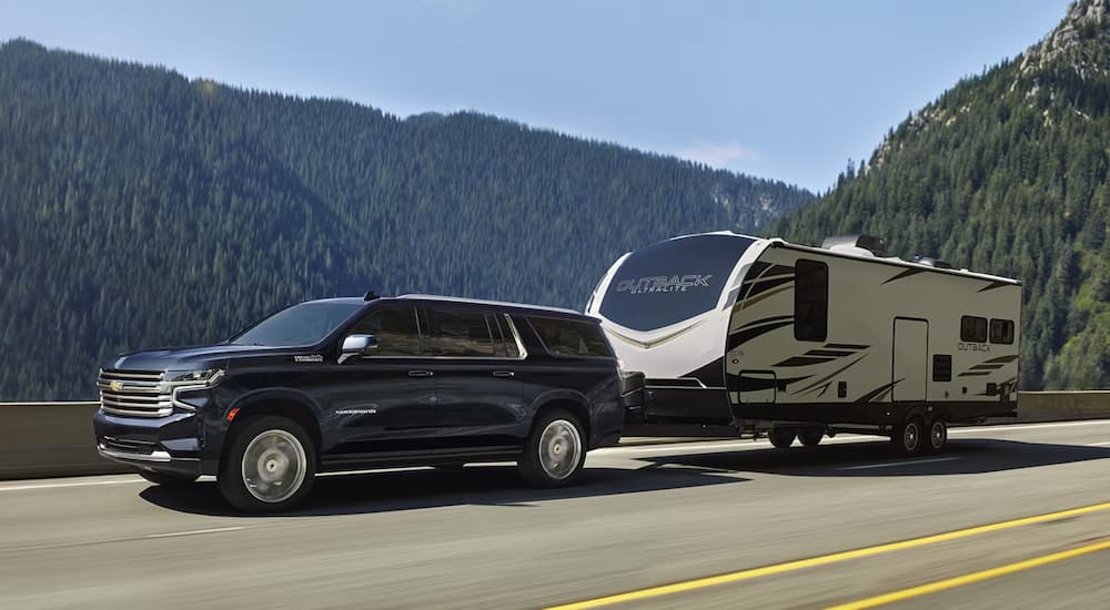A blue 2024 Chevy Suburban High Country towing a large camper on a mountain highway.
