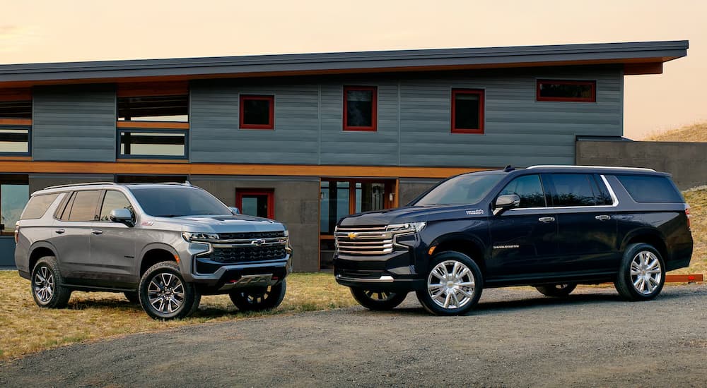 A silver 2024 Chevy Suburban Z71 and a blue 2024 Chevy Suburban High Country parked in front of a house.