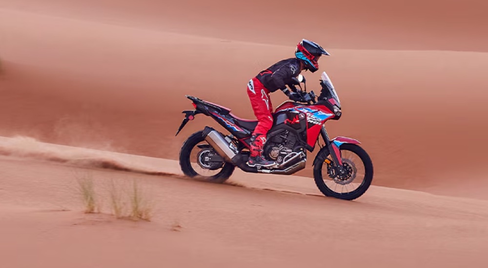 A rider in colorful gear riding a matching 2024 Honda Africa Twin DCT down a sand dune.