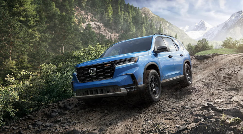 A blue 2025 Honda Pilot TrailSport is shown off-roading on a rocky dirt road.