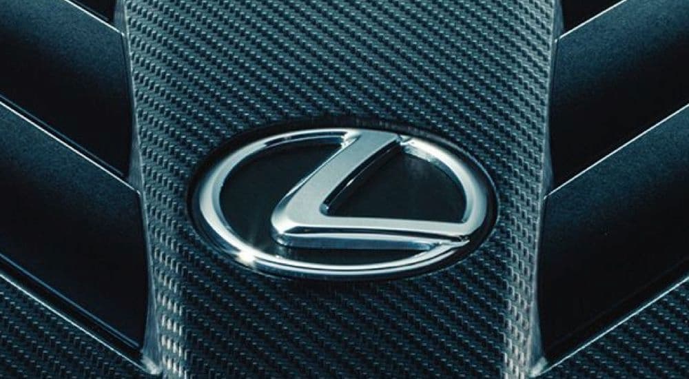 A close-up of the silver Lexus logo mounted on a black carbon fiber grille at a used Lexus dealer.