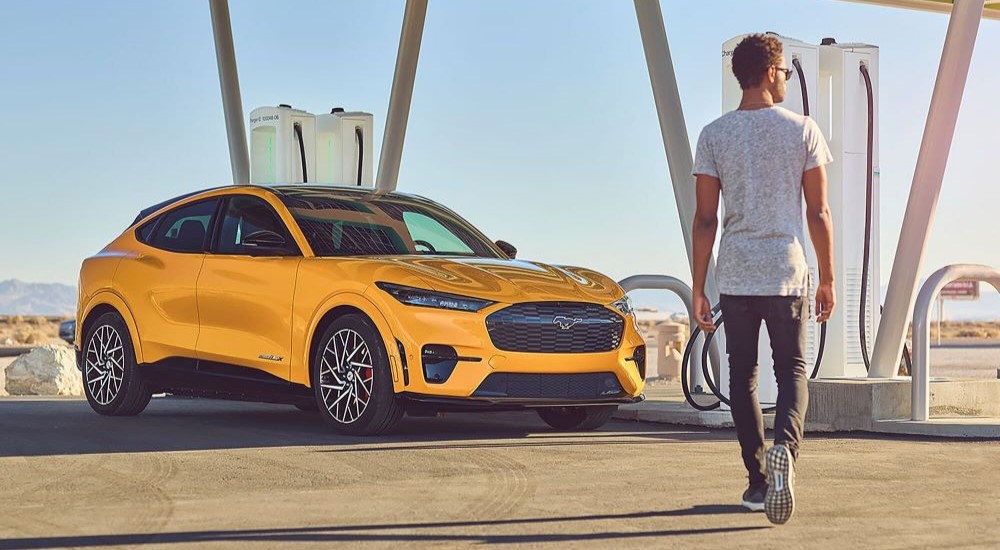 A man walking towards a yellow 2024 Ford Mustang Mach-E GT parked at a charging station after visiting a Ford dealer.