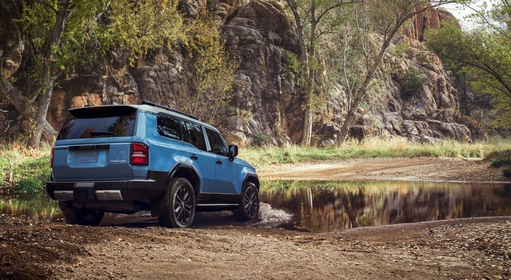 A blue 2024 Toyota Land Cruiser is shown driving off-road near water after visiting a Toyota dealer.