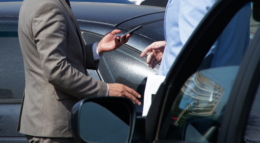 A car salesman is shown taking to a customer at a dealership lot with used cars for sale.