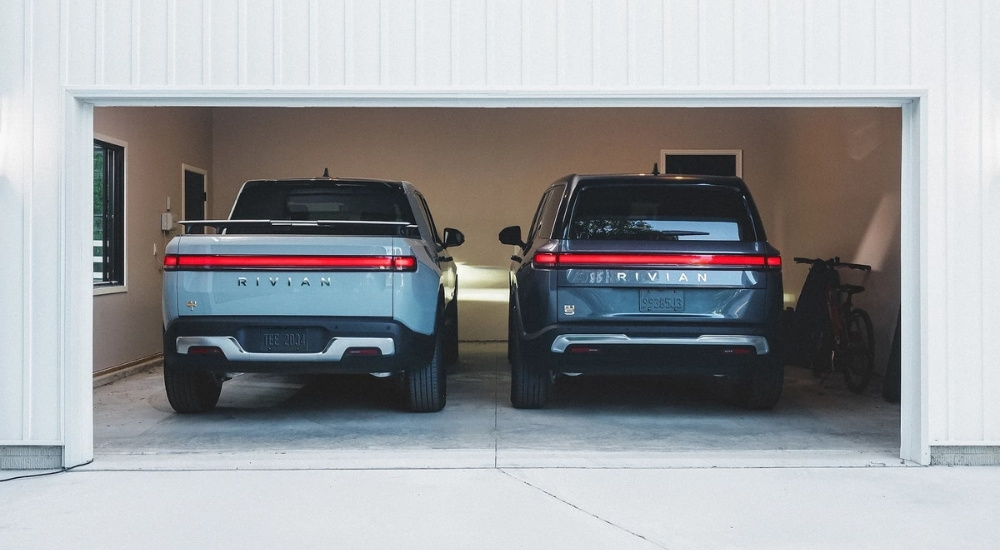 A light blue 2024 Rivian R1T and a dark blue Rivian R1S are shown in a garage.
