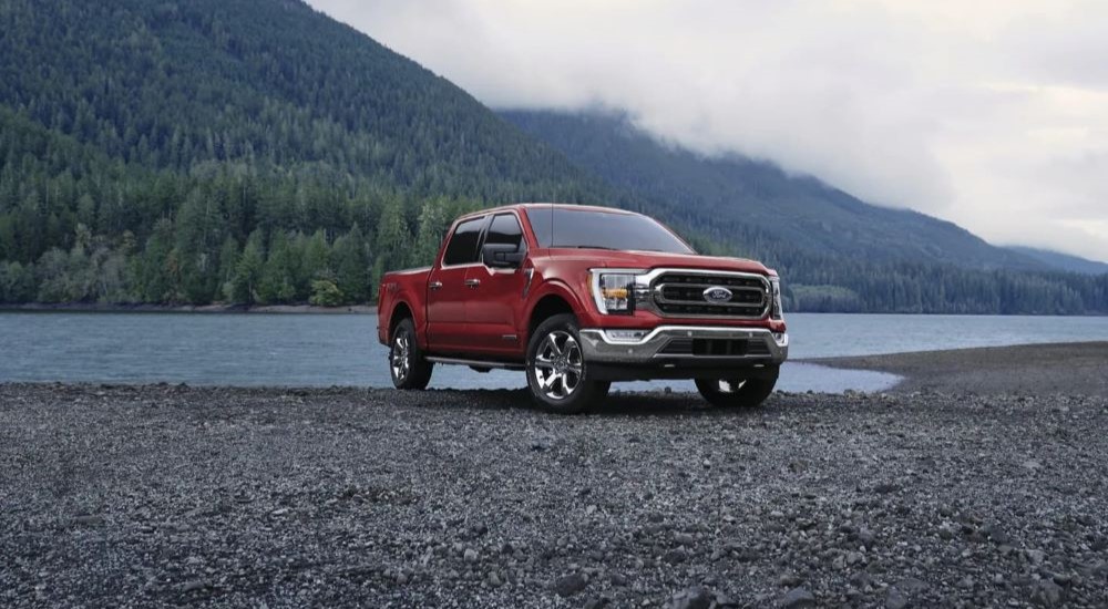 Red 2023 Ford F-150 parked near the water of a river with trees and clouds behind it.