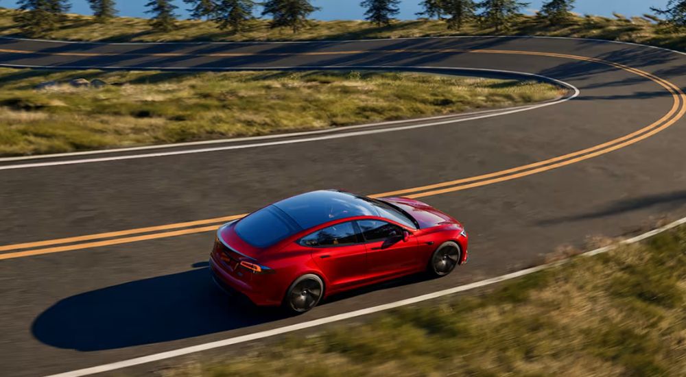 Red 2026 Tesla Model S driving on a tree lined road near a lake
